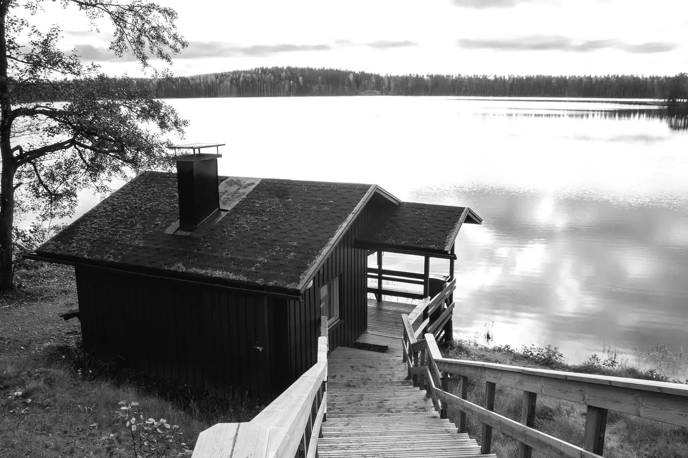 A dark-stained sauna cabin at the end of a wooden boardwalk running down to a still lake, with forest on the far shore.