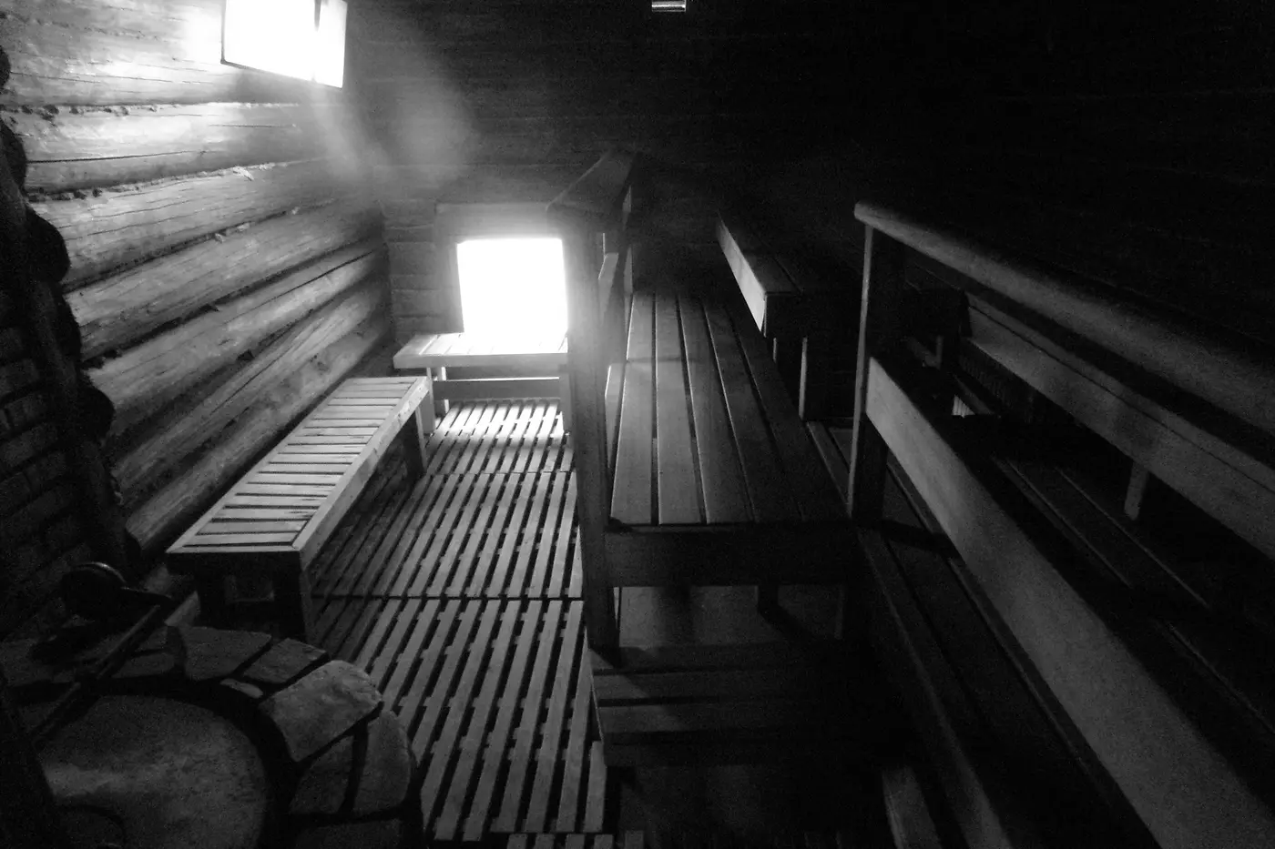 The interior of a log sauna lit by one small window, benches and duckboards in low light with steam hanging in the air.