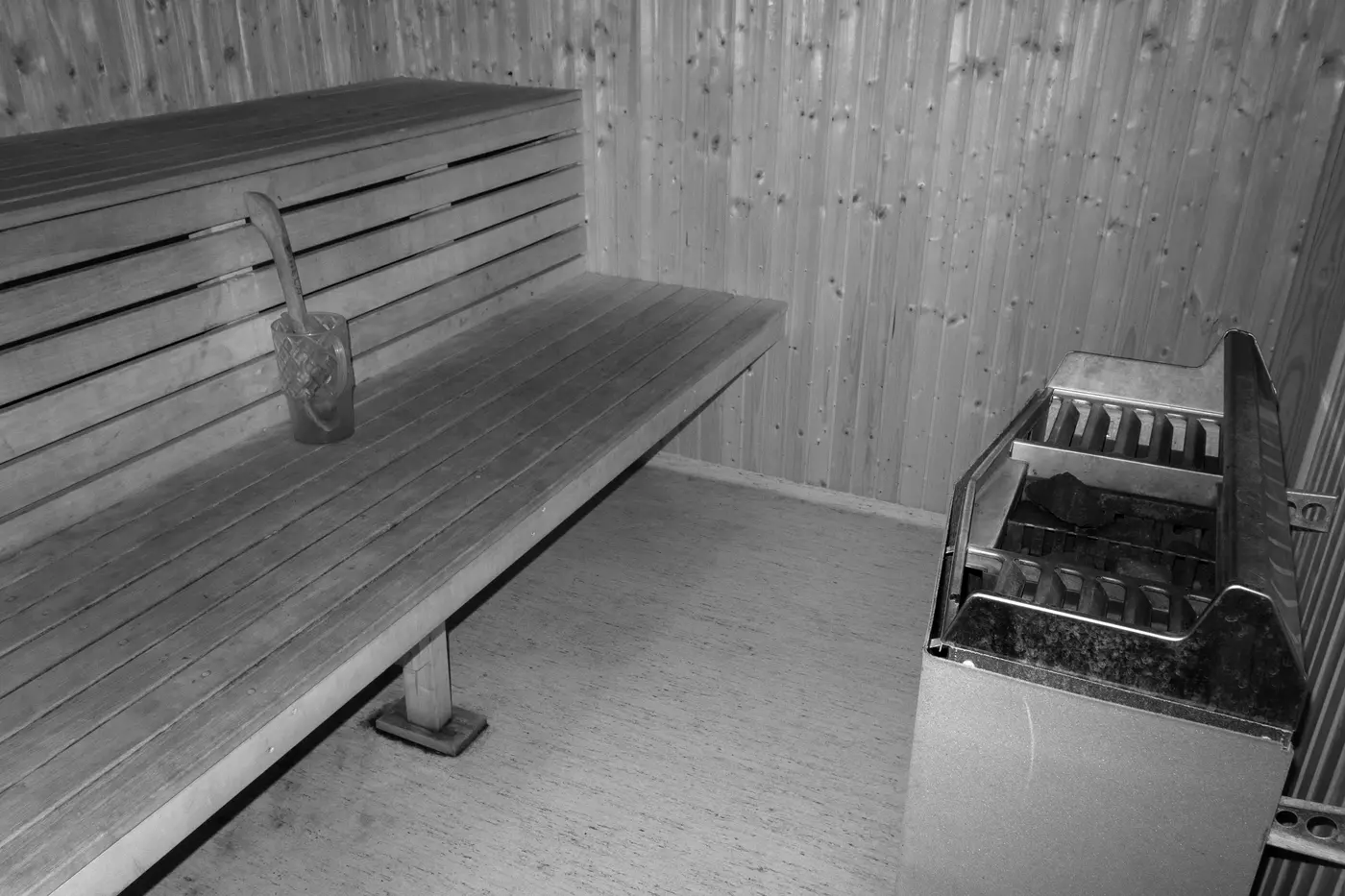 A pale wooden bench running the length of a small sauna, with a stainless electric heater standing beside it and a wooden dipper hooked over the backrest.