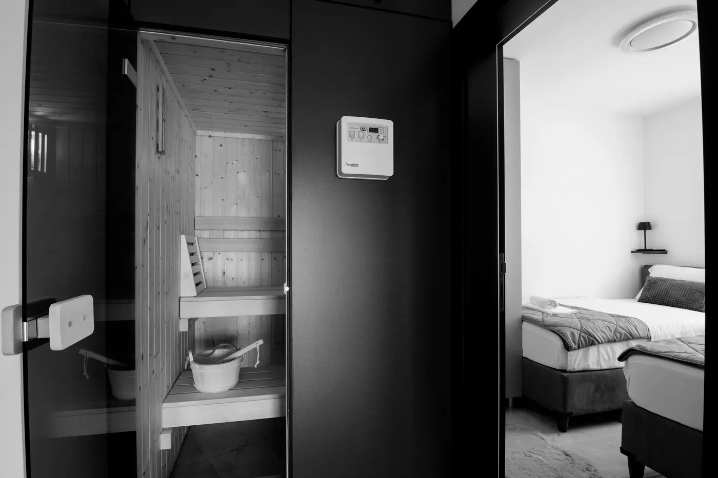 A compact glass-doored sauna built into a modern apartment beside a bedroom, with a wall-mounted control unit on the panel between them.