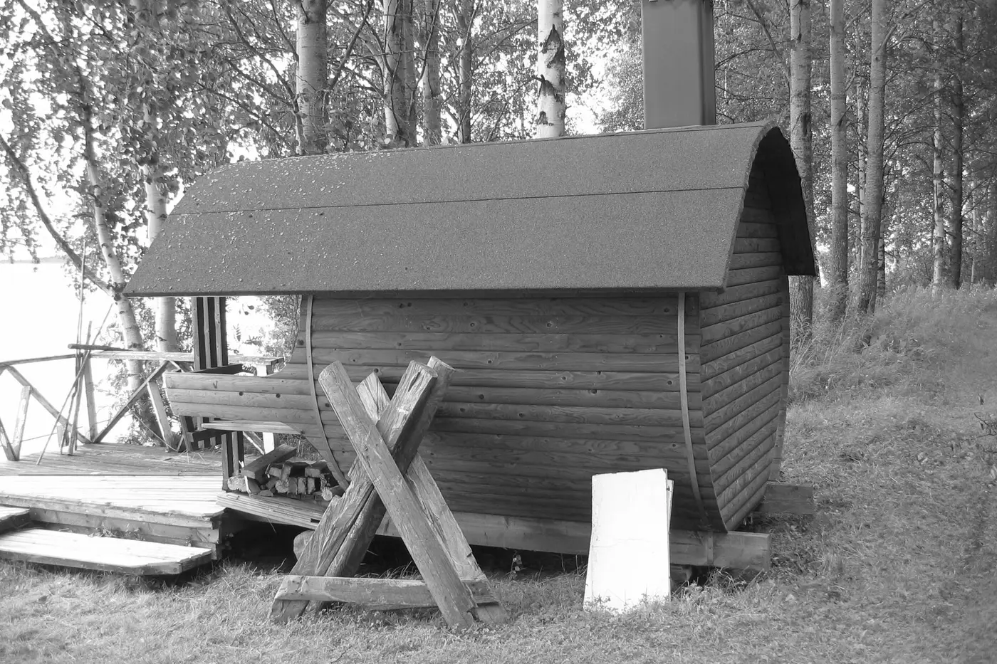 A barrel sauna built from stacked timber on a lakeshore, roofed in felt, with cut firewood stacked beneath the eaves and birch trees behind.