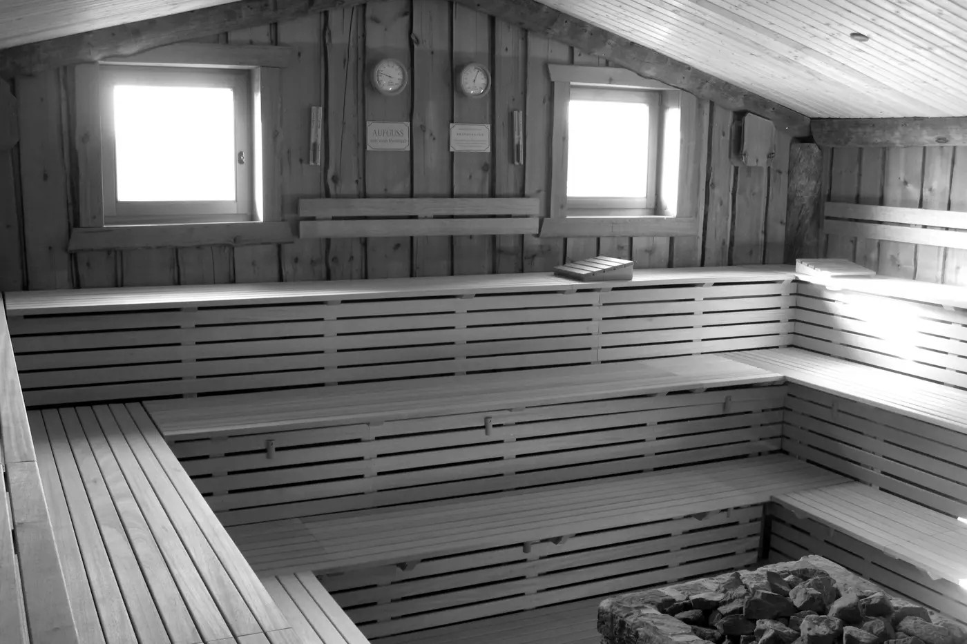 A large public sauna lined entirely in pale timber, with tiered slatted benches running around three walls beneath two small windows.